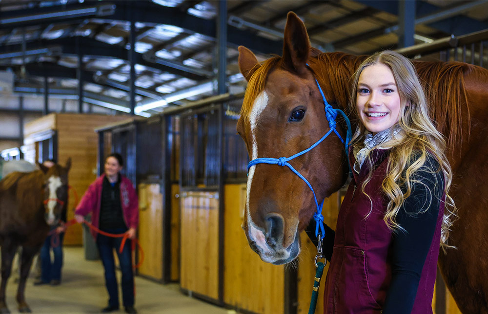 A smiling student holds the reigns of a brown horse, behind her another student walks a horse through the barn full of stables.